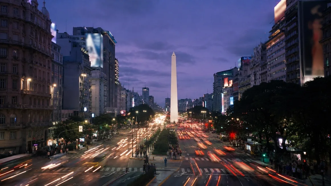 The Obelisco of Buenos Aires at dusk, seen down a wide avenue streaked with traffic light-trails under a violet sky