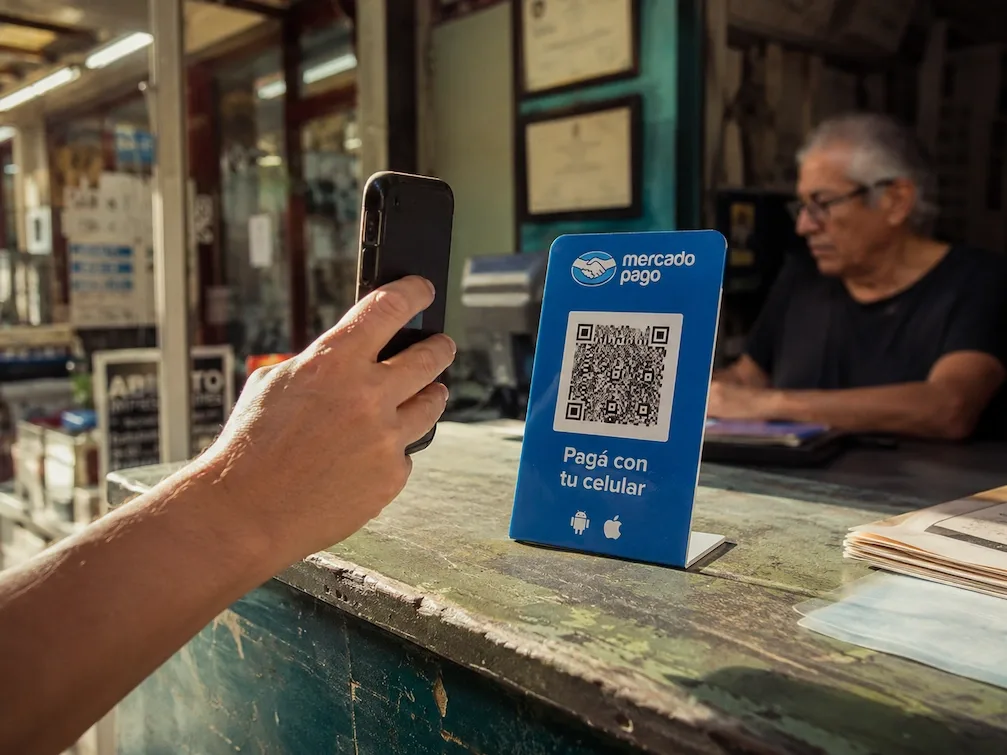 A hand holds a phone up to the blue Mercado Pago QR standee on a Buenos Aires shop counter