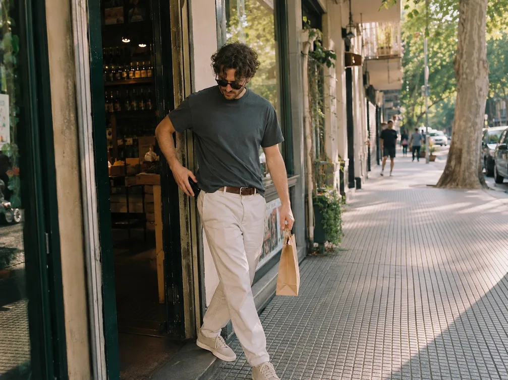 A shopper steps out onto a sunlit Palermo sidewalk with a paper bag, pocketing their phone