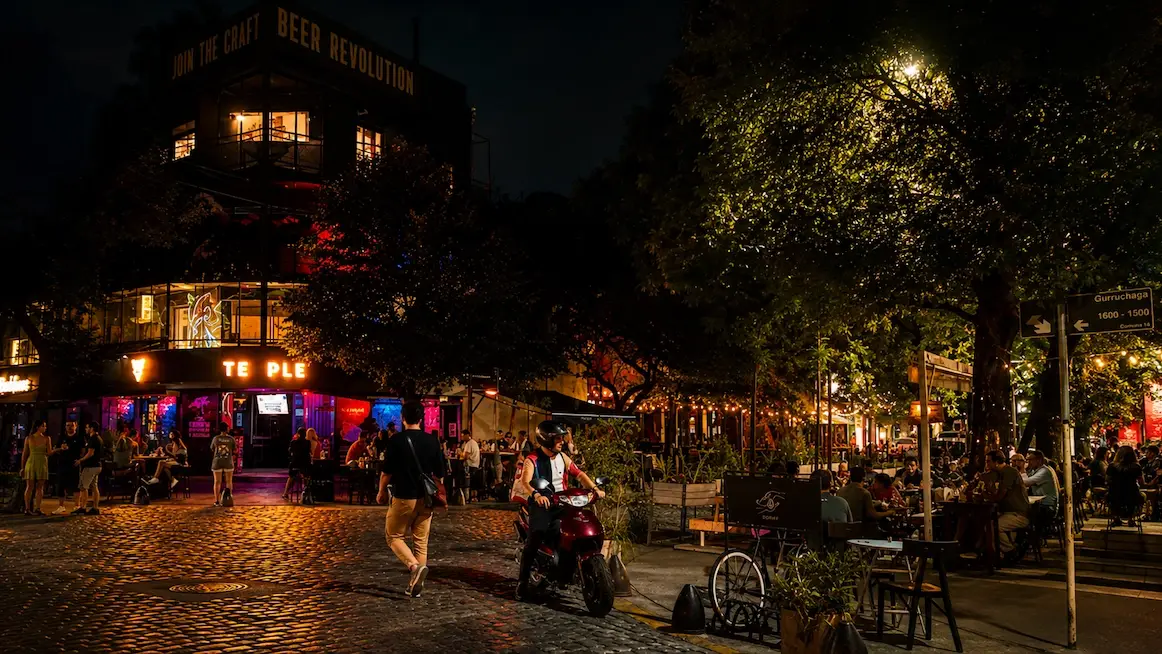 A lively Palermo Soho corner at night, a cobblestone street lined with string-lit sidewalk bars and a lit corner building topped with a craft-beer sign, people at outdoor tables and a motorbike crossing the intersection