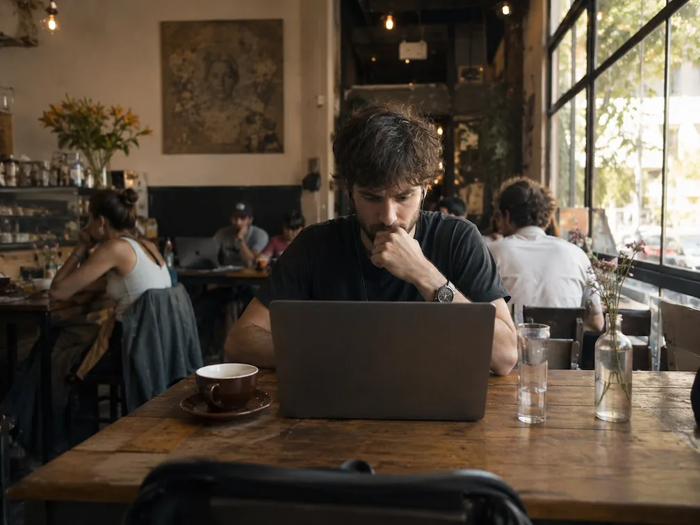 A person works on a laptop at a wooden table in a busy, light-filled Buenos Aires cafe