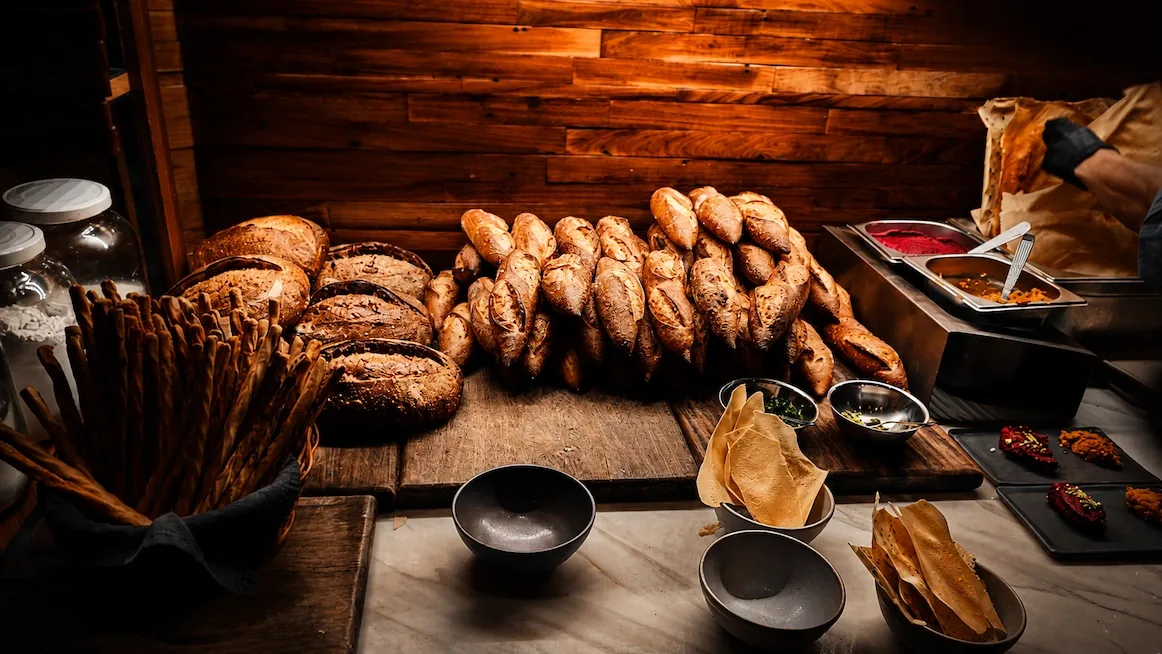 A warmly lit counter piled with house-baked breads — twisted baguettes and dark round sourdough loaves on a wooden board, a basket of grissini, and small bowls of dips and crisps on marble.
