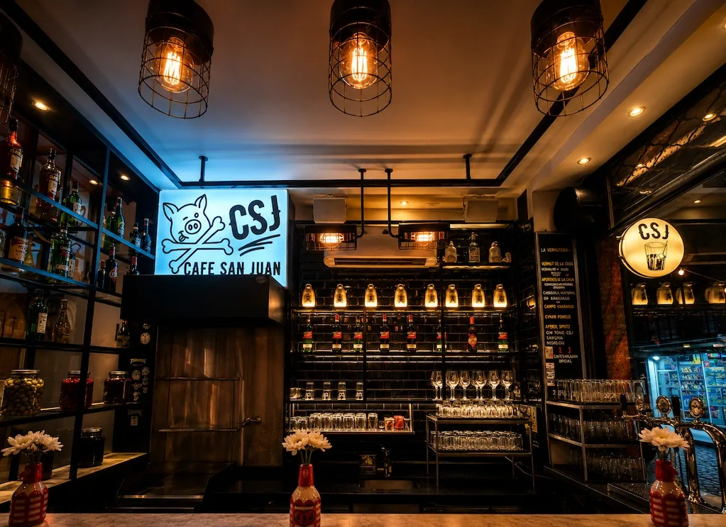 The Café San Juan bar, its pig-and-crossbones sign lit over shelves of bottles, rows of wine glasses, and caged filament bulbs, with flowers on the marble counter.