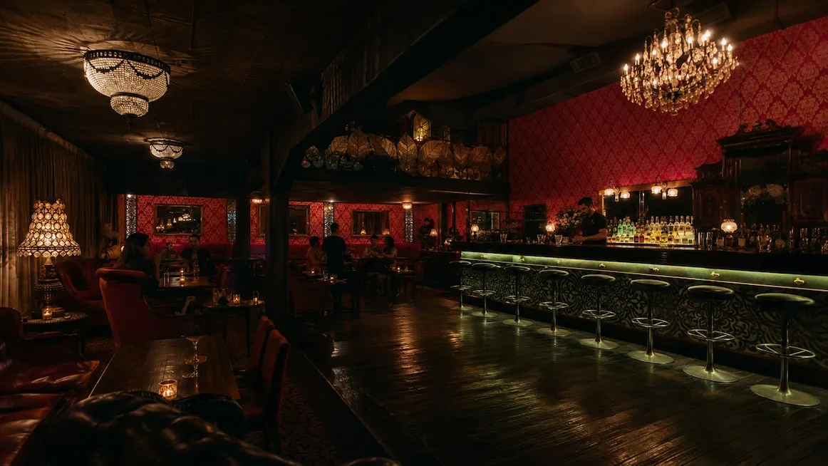 A chandelier-lit speakeasy with deep red damask walls — a long bar glowing green along its base with chrome stools, and tufted leather armchairs around candlelit tables where guests sit.