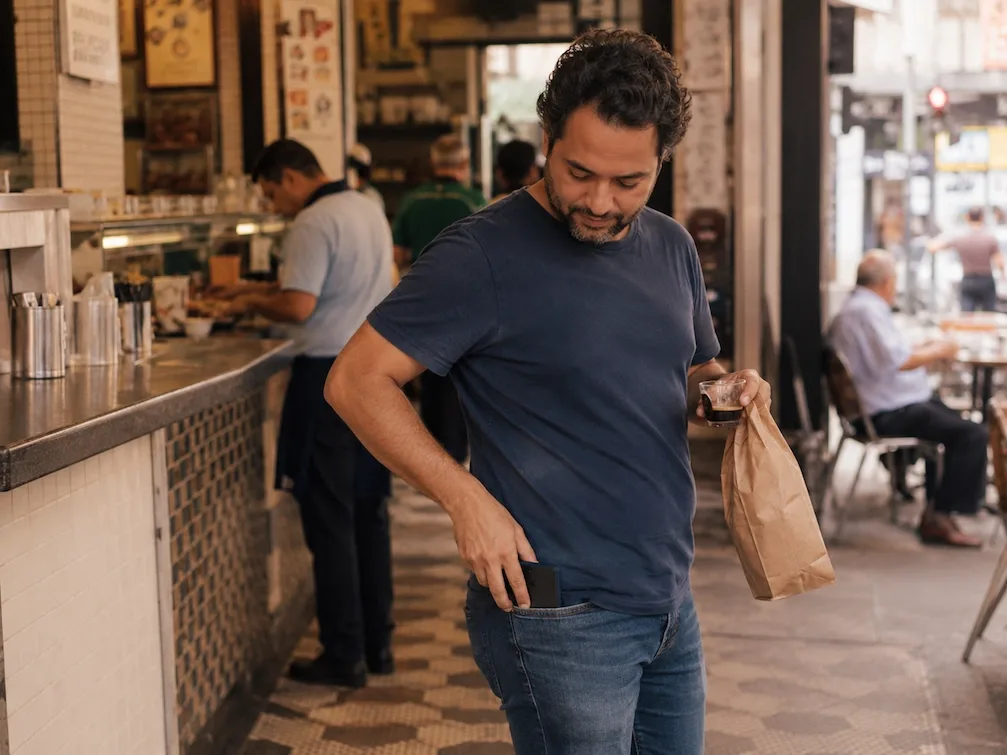 A customer steps away from a Sao Paulo lunch counter with a coffee and a paper bag, pocketing their phone