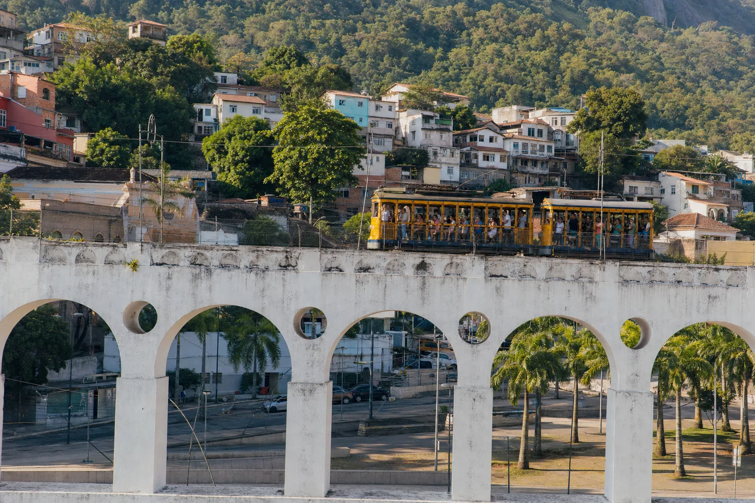 Santa Teresa's yellow bonde tram crossing the Arcos da Lapa aqueduct in daylight, passengers hanging from the open sides