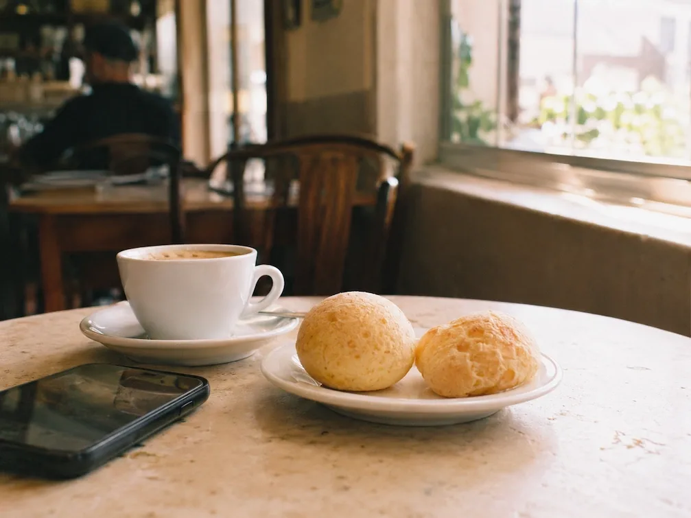 A cup of coffee and a plate of pão de queijo on a café table by a sunlit São Paulo window, a phone resting beside them