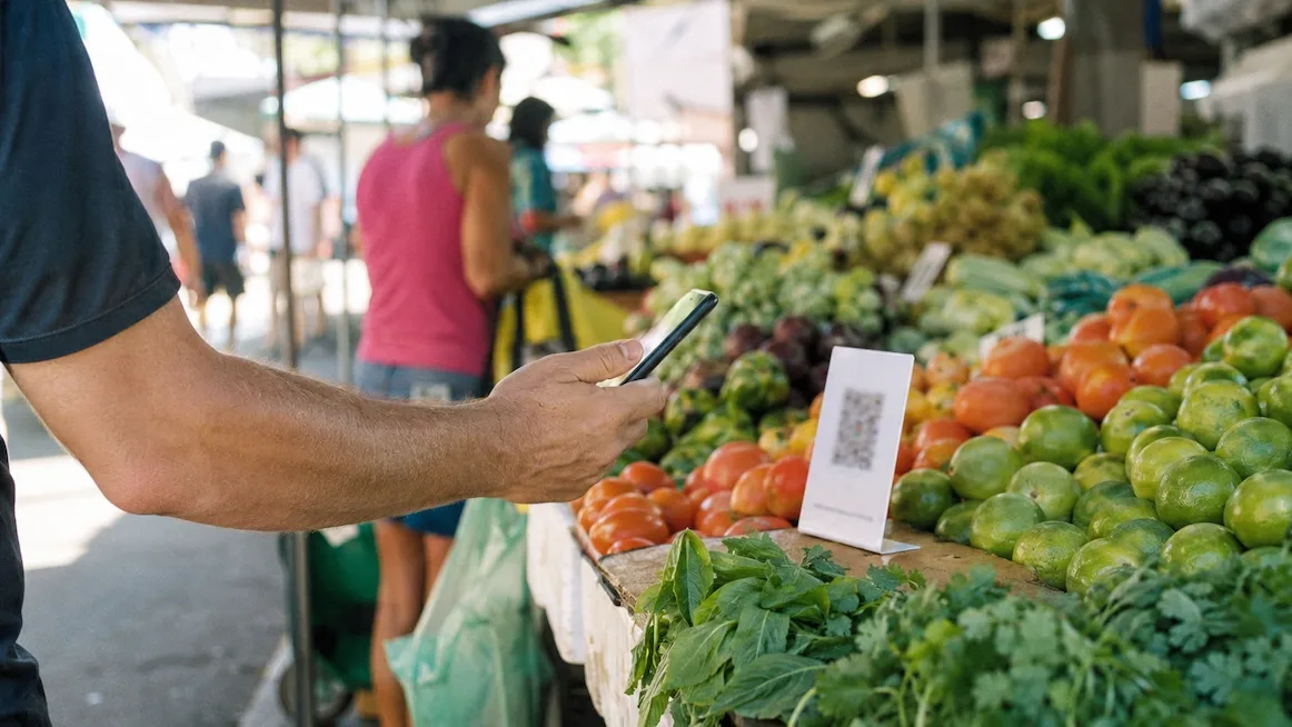 A customer holds a phone up to a plain QR-code standee at a São Paulo street-market produce stall piled with tomatoes, limes and greens