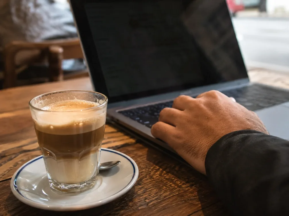 A coffee in a glass beside an open laptop on a wooden cafe table, a hand on the keys
