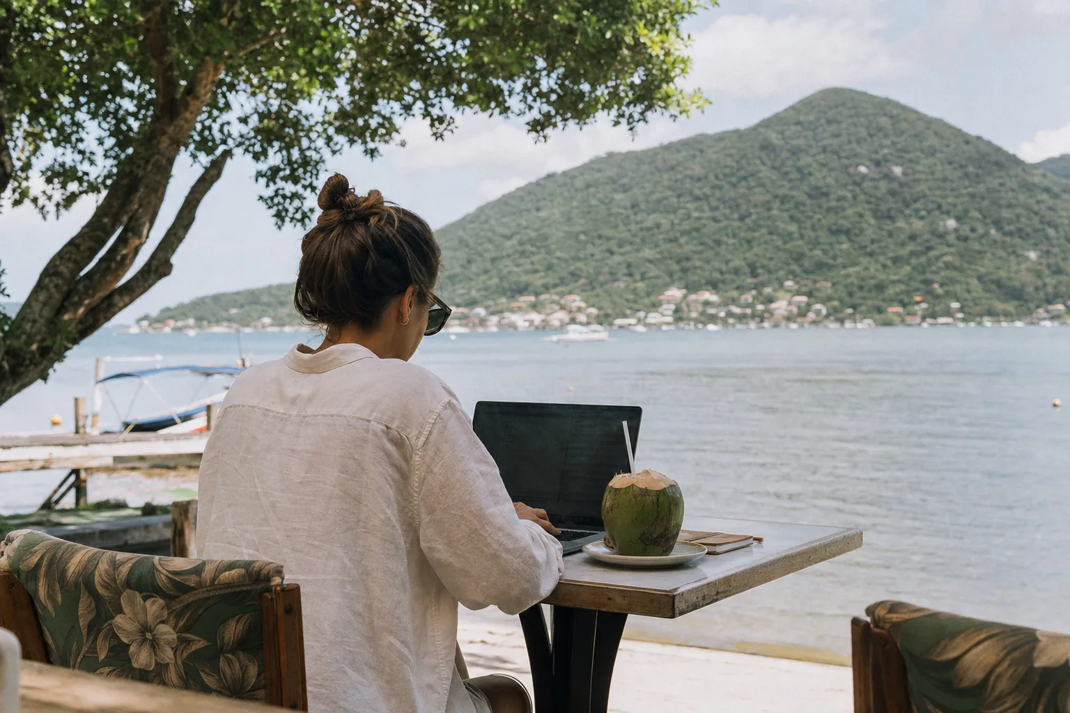 A person working on a laptop at an outdoor cafe table near the Lagoa da Conceicao waterfront in Florianopolis