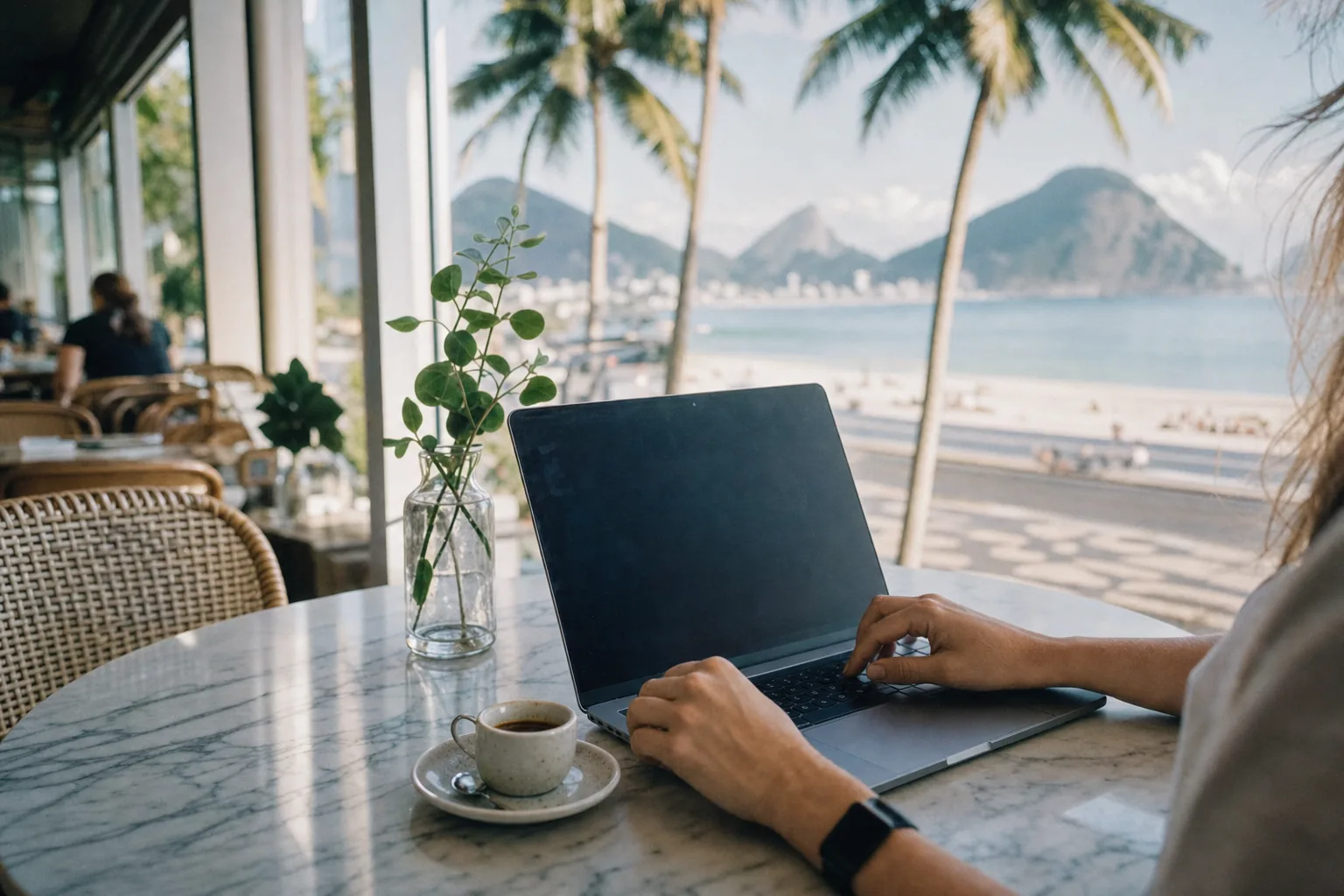 A laptop and a small cup of coffee on a marble-top table at a Rio de Janeiro café, morning light through large windows