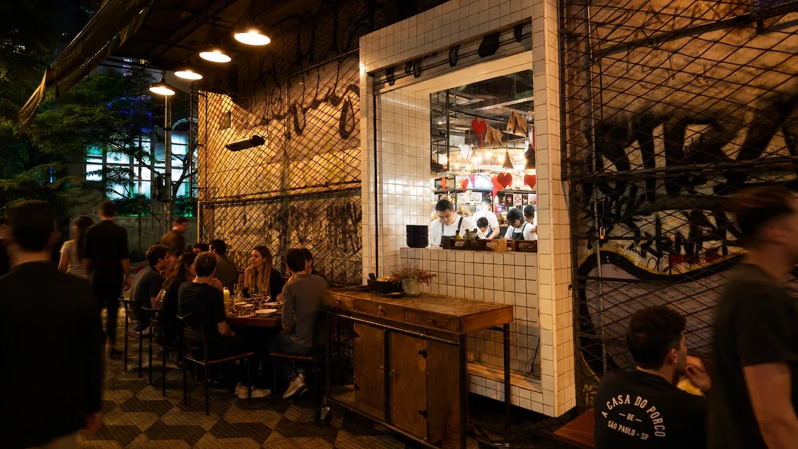 The night terrace at A Casa do Porco — diners at tables against a graffitied wall, a white-tiled window onto the open kitchen where aproned cooks work, and a guest in a branded A Casa do Porco shirt in the foreground.