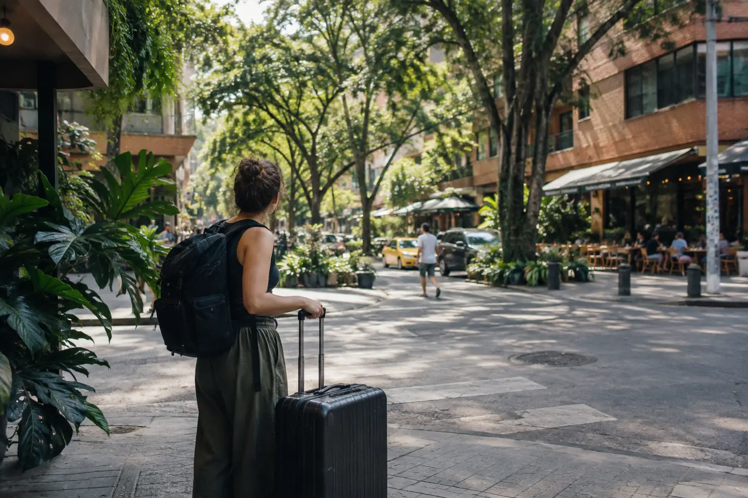 A traveler pauses with a suitcase on a tree-shaded street corner, brick buildings and sidewalk tables in Laureles