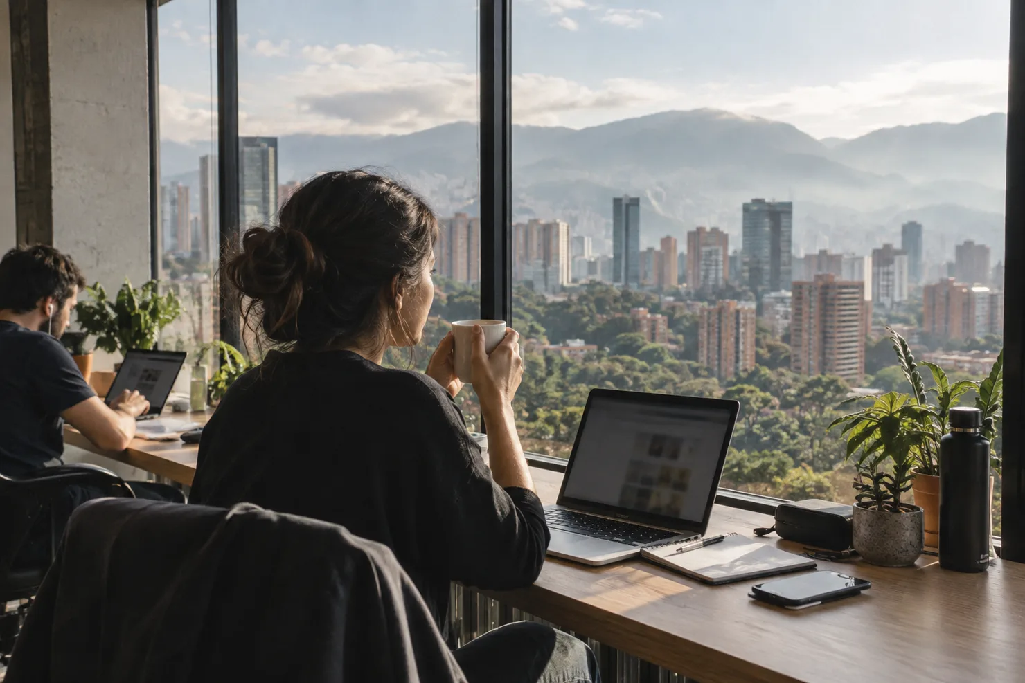 A coworking desk by a tall window looking out over El Poblado's towers and tree canopy, a laptop, a notebook and a phone on the desk