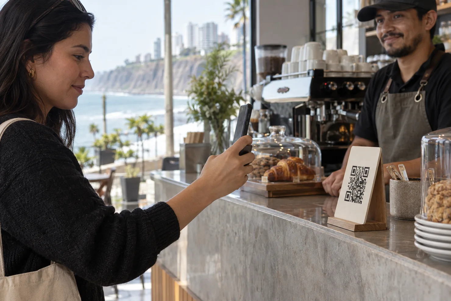 A customer holding a phone up to a QR standee at a Miraflores café counter, the screen turned away, the sea behind the window