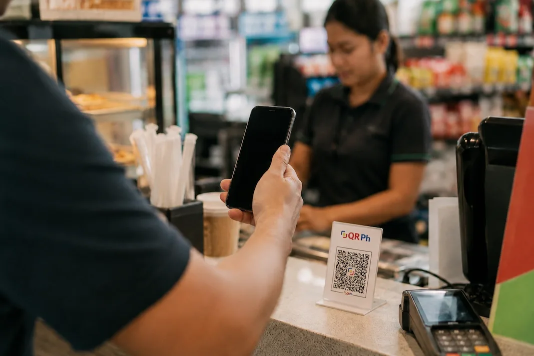 A customer holds a phone up to a QR-code standee at a Manila convenience-store counter, a cashier and a card terminal beside it