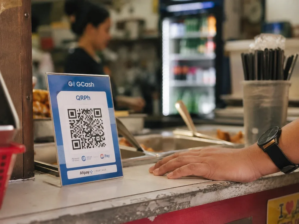 A GCash QRPh payment standee on a Manila food-stall counter, a customer hand resting beside it