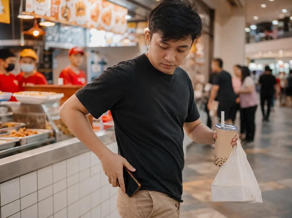 A young man leaves a Manila food court with a milk tea and a takeout bag, pocketing his phone