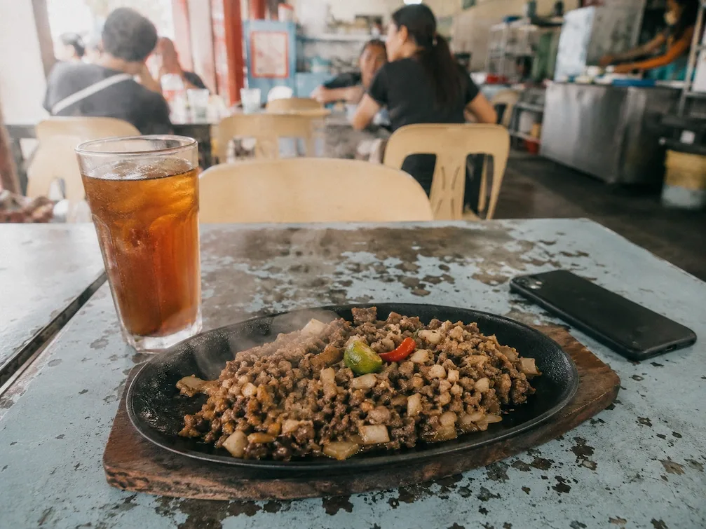 A sizzling plate of sisig with calamansi and chili, an iced tea and a phone face-down on a worn eatery table