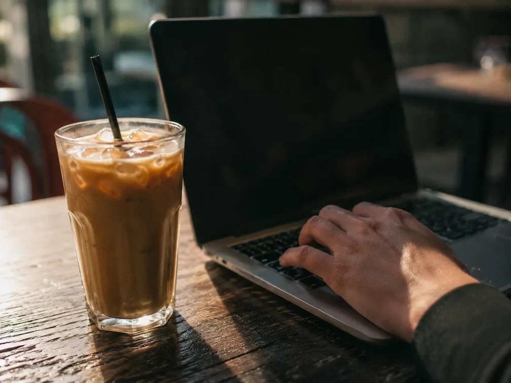 A tall iced coffee beside an open laptop on a cafe table, a hand on the keys