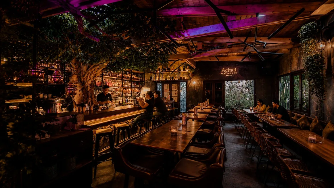 The jungle-like interior of Agimat Foraging Bar — a live tree and hanging greenery under magenta light, a backlit wall of bottles behind the bar, long communal tables, and the lit Agimat sign on the back wall.