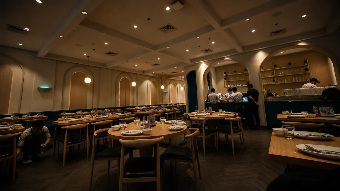 The dining room at Manam — round wooden tables set with bowls and glasses under a coffered ceiling and arched wood-paneled niches, staff at the service counter at the back.