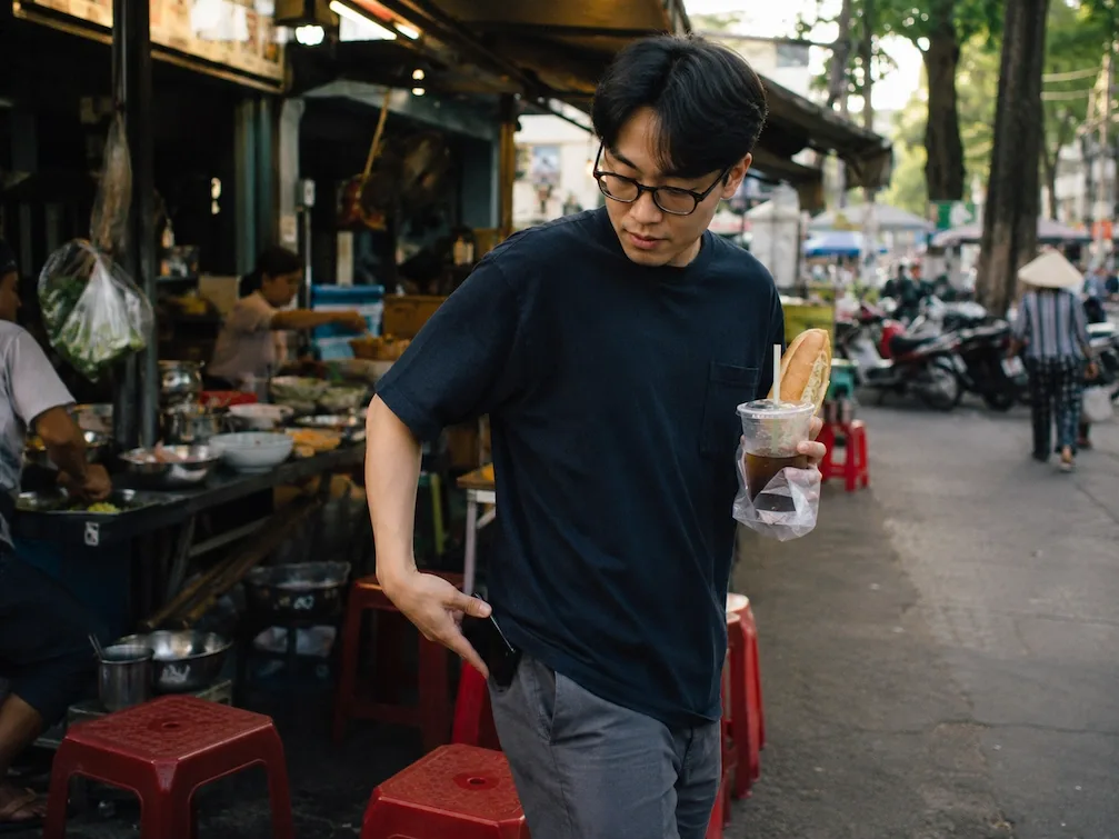 A customer leaves a Ho Chi Minh City street-food stall with a banh mi and iced coffee, pocketing his phone