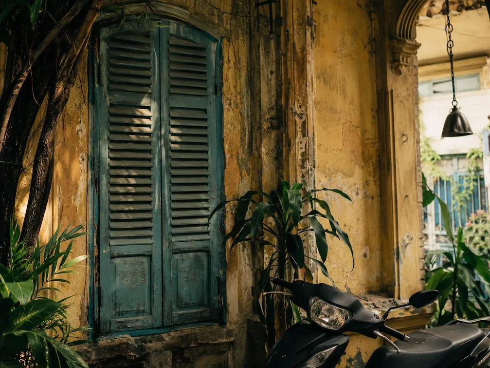 A close view of a weathered French-colonial shophouse with green louvered shutters and a parked motorbike