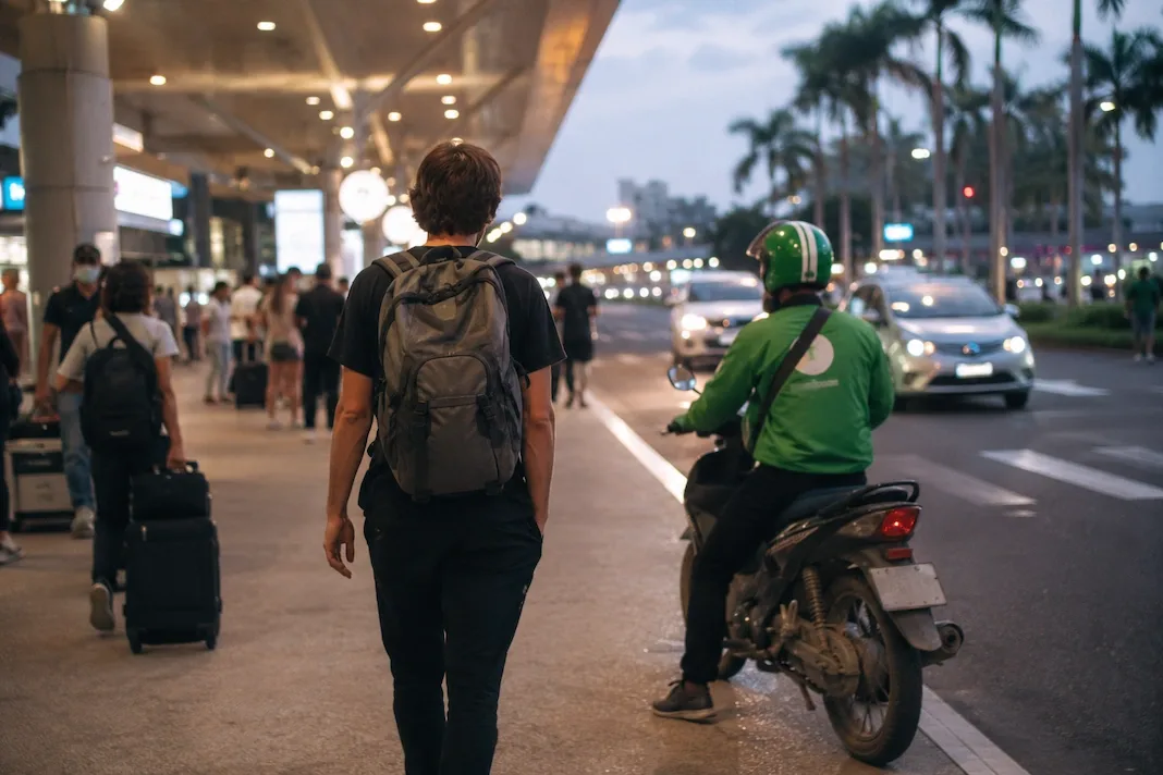 A backpacker seen from behind walks an airport arrivals curb toward a waiting green-jacketed motorbike-taxi driver, palms and terminal lights beyond