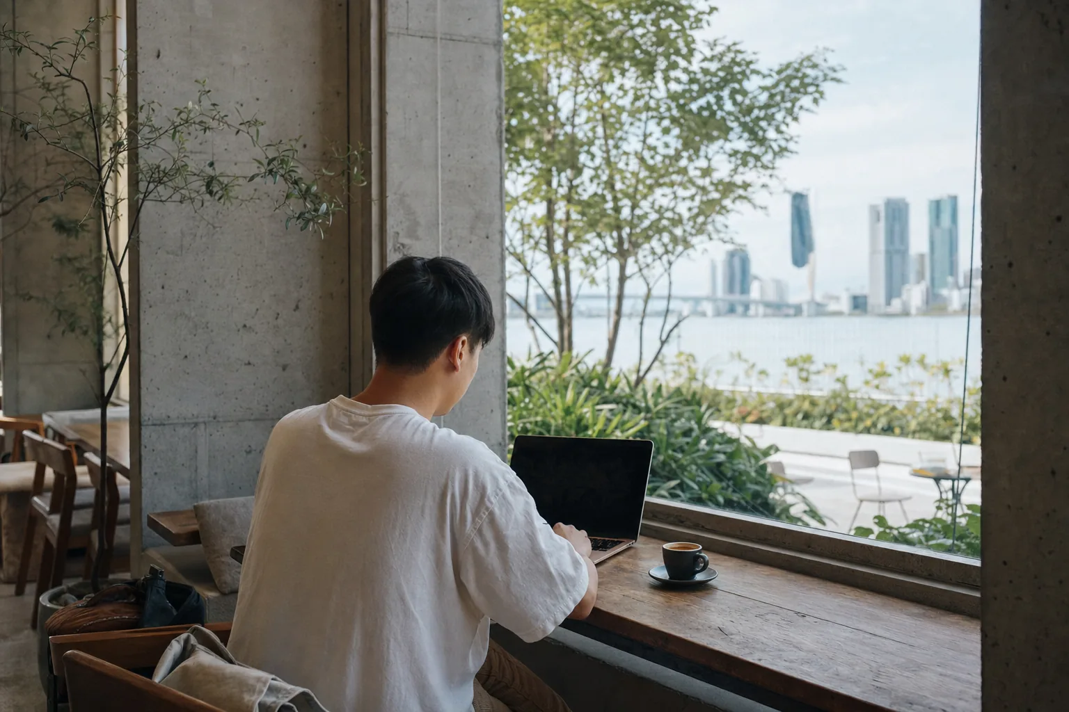 A person works on a laptop at a window-side table in a minimalist concrete-and-glass Da Nang cafe, an espresso beside the keyboard