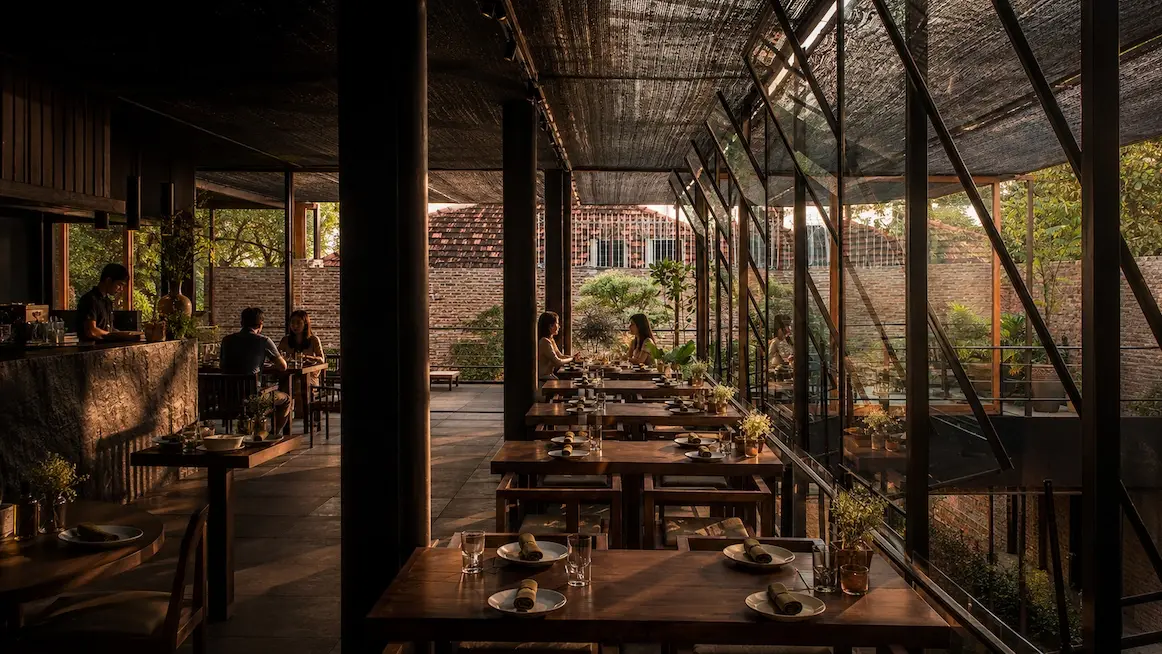 A Pizza 4P's dining room — long wooden tables set for dinner under woven-bamboo shade, angled glass walls opening onto a bare-brick courtyard with greenery, and a stone bar to one side.