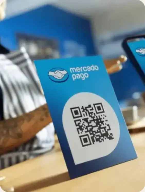 A blue Mercado Pago QR code card on a cafe counter, with a phone scanning it beside it.