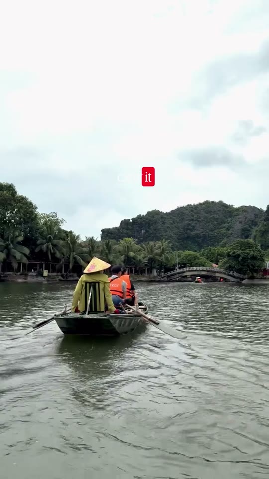 A wooden boat with rowers in conical hats glides down a calm green river past palm-covered karst hills in Vietnam.